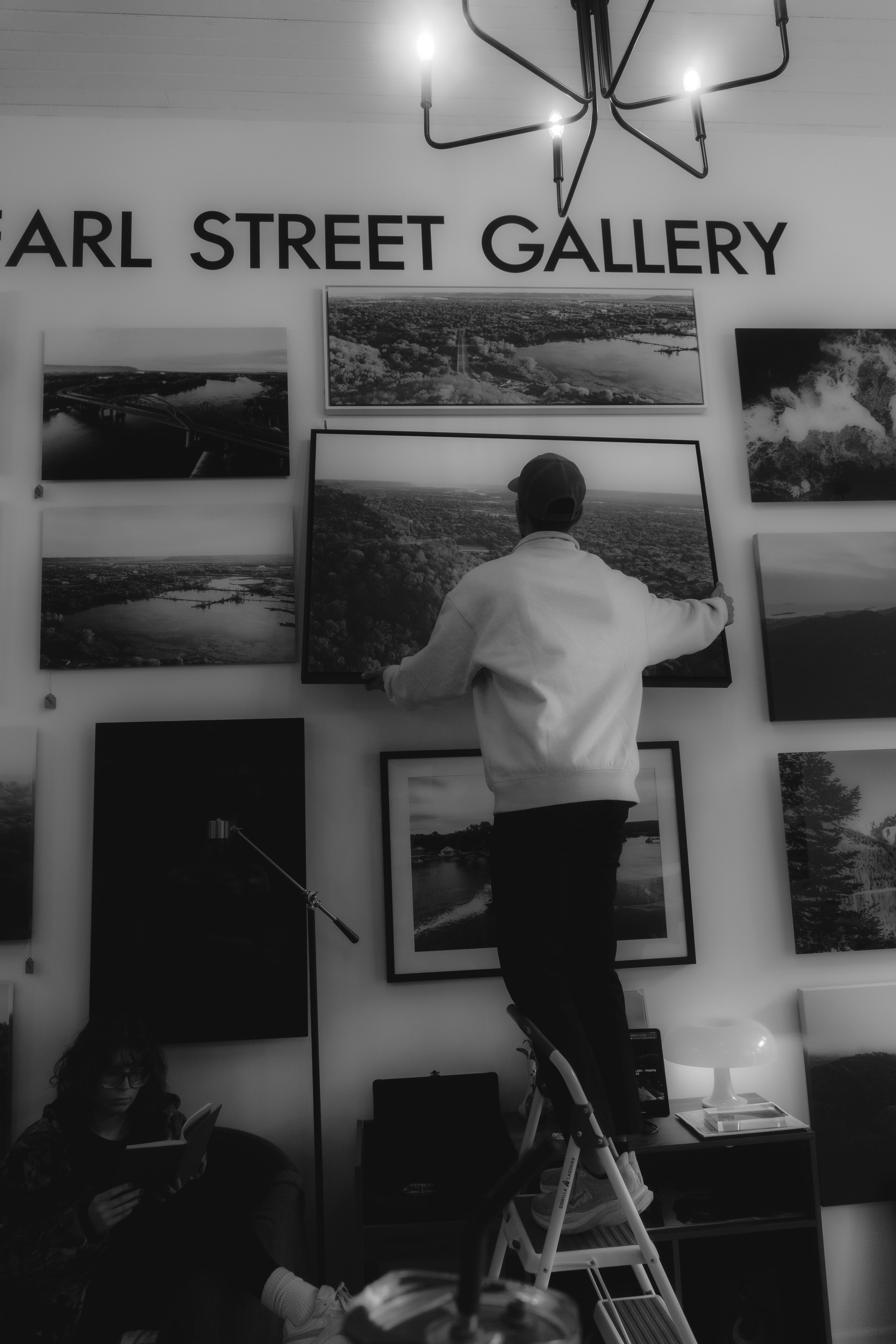 Person on a ladder hanging artwork in a gallery with 'Pearl Street Gallery' sign.